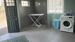 Ein Waschraum mit Waschmaschine, Wäscheständer, einem kleinen Hocker und einem Fenster mit Spitzenvorhängen.
