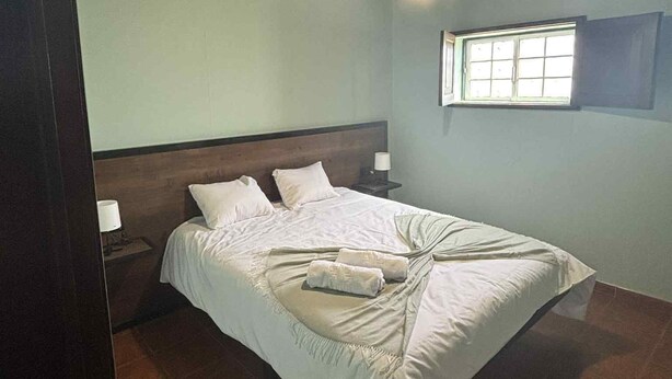 Ein Schlafzimmer mit Holzbalkendecke, einem Einzelbett mit weißer Bettwäsche, einem Nachttisch mit Lampe und einem Fenster mit Jalousien.
