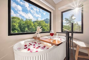 A spa tub with rose petals, a glass of red wine, and a view of a mountainous landscape through large windows. - Riverside Hot Tub, Cedar Sauna & Arcade Games (Hunter)