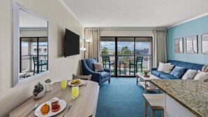 Ein Hotelzimmer mit Balkon, einer Essecke mit Frühstück, einer Couch und einem an der Wand befestigten Fernseher.