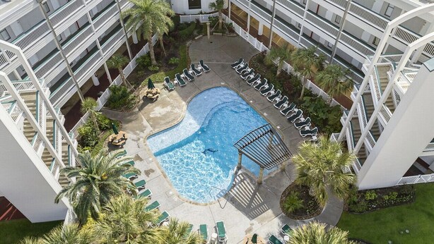 Vue aérienne d’un hôtel doté d’une piscine centrale, entourée de chaises longues et de palmiers.