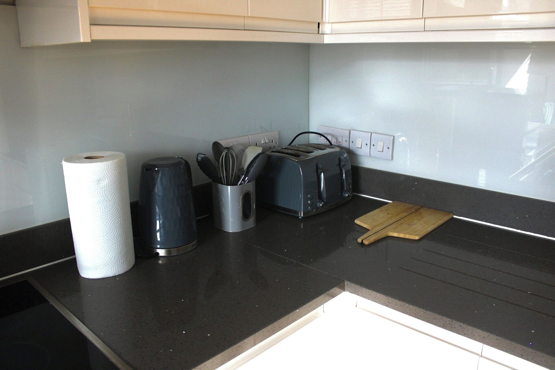 Cozinha com bancada preta, torradeira, cafeteira e tábua de corte de madeira.
