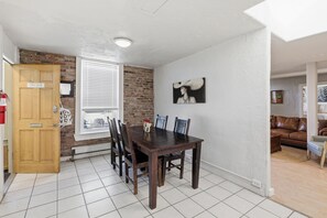 A dining area with a wooden table and chairs, a brick wall, a white ceiling, and a window with blinds.