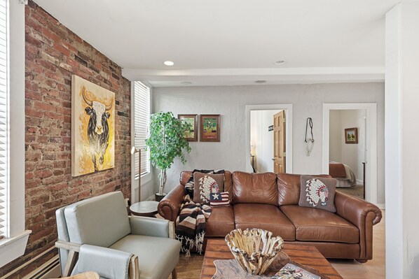 A living room with a brown leather sofa, a light grey armchair, and a wooden coffee table. There is a large plant, a framed picture on the wall, and a brick wall.
