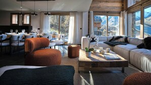Una sala moderna con sofá, reposapiés y mesa de centro, un comedor con sillas y mesa, y ventanas amplias con vistas a la montaña.