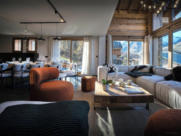 A modern living room with a sofa, ottoman, and coffee table, a dining area with chairs and a table, and large windows offering a mountain view. - Chalet Fango - Splendid modern chalet with sauna and hot tub - OVO Network (Les Belleville)