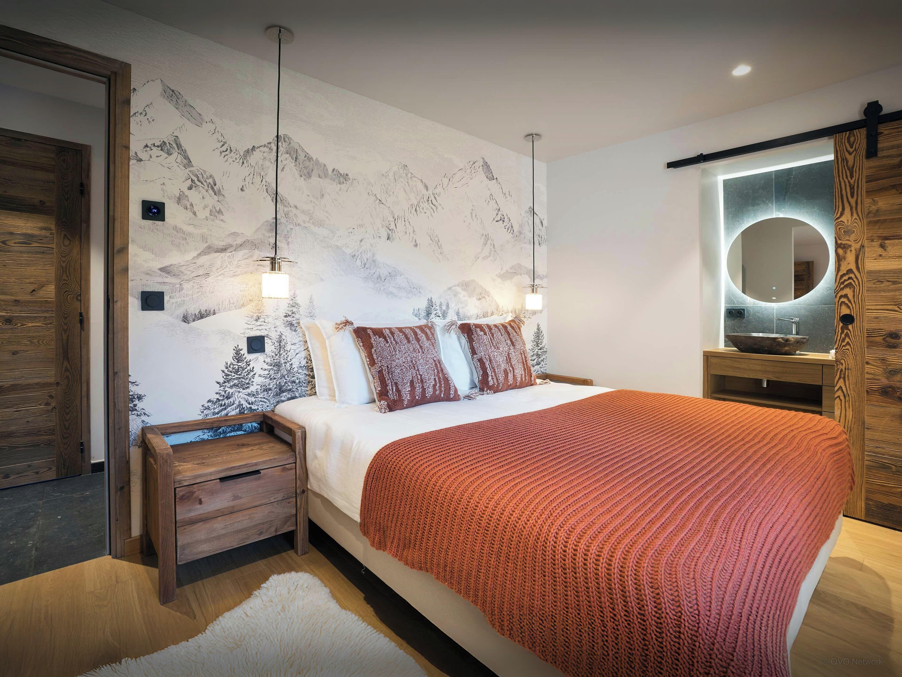 Un dormitorio moderno con una cama grande, mesitas de noche de madera, un armario de madera, un espejo redondo y una pared con un mural de montañas.
