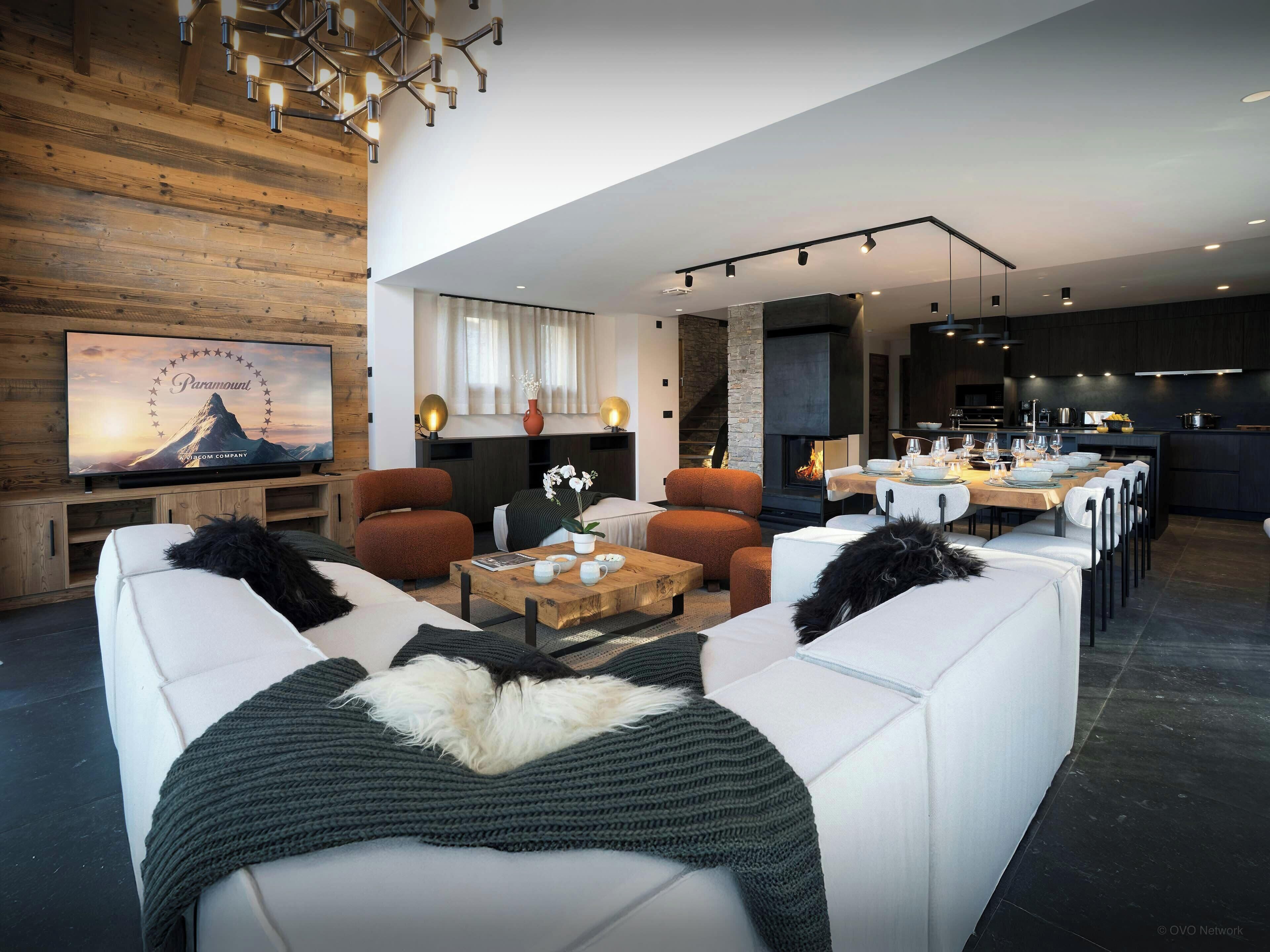Ein modernes Wohnzimmer mit weißen Sofas, einer Holzvertäfelung, einem Kamin und einem Essbereich, im Hintergrund befindet sich eine Küche.