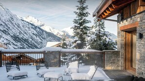 Un área de estar al aire libre cubierta de nieve con un jacuzzi, paredes de piedra y un paisaje de montañas al fondo.