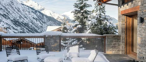 A snow-covered outdoor seating area with a hot tub, stone walls, and a mountain backdrop.