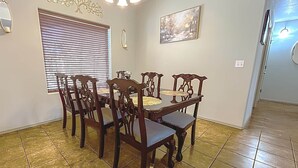 A dining area with a wooden table and chairs, a window with blinds, a chandelier, and a painting on the wall. - Luxury Vacation Rental (Las Cruces)