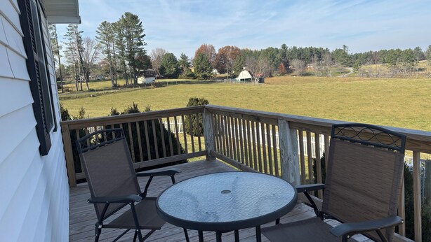 A wooden deck with a round table and two chairs, overlooking a grassy field and a white building.