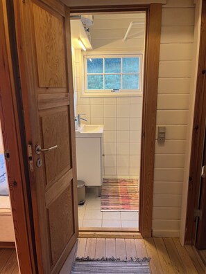 Puerta de madera que da a un baño con ventana, lavamanos y cubo de basura.