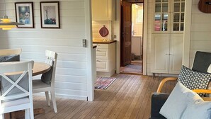 A cozy living room with a wooden floor, a dining table with chairs, a sofa with cushions, and a kitchen visible through an open doorway. - Log cabin on Fanø near beach and nature (Fanø)