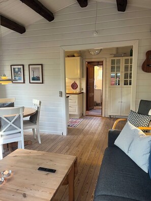 Ein gemütliches Wohnzimmer mit Holzboden, einem Esstisch mit Stühlen, einer Couch mit Kissen und einer durchgehenden Tür erkennbaren Küche.