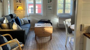 A cozy living room with a wooden coffee table, a sofa, and a chair. There are two windows with curtains, a lamp, and wall art. - Log cabin on Fanø near beach and nature (Fanø)