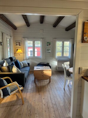 Ein gemütliches Wohnzimmer mit einem Holztisch, einer Couch und einem Sessel. Zwei Fenster mit Vorhängen, eine Lampe und Wandbilder sind zu sehen.