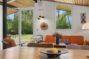 Ein Innenraum mit Esstisch, Stühlen, einer Couch und Blick auf einen Garten durch große Fenster.