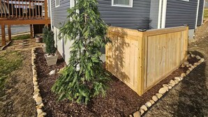 A two-story house with a dark siding exterior, a small front yard with a wooden deck, and a young coniferous tree. - ★ Superhost Rated ★ Lily of the Valley Retreat - Steps to Downtown (Kingsport)