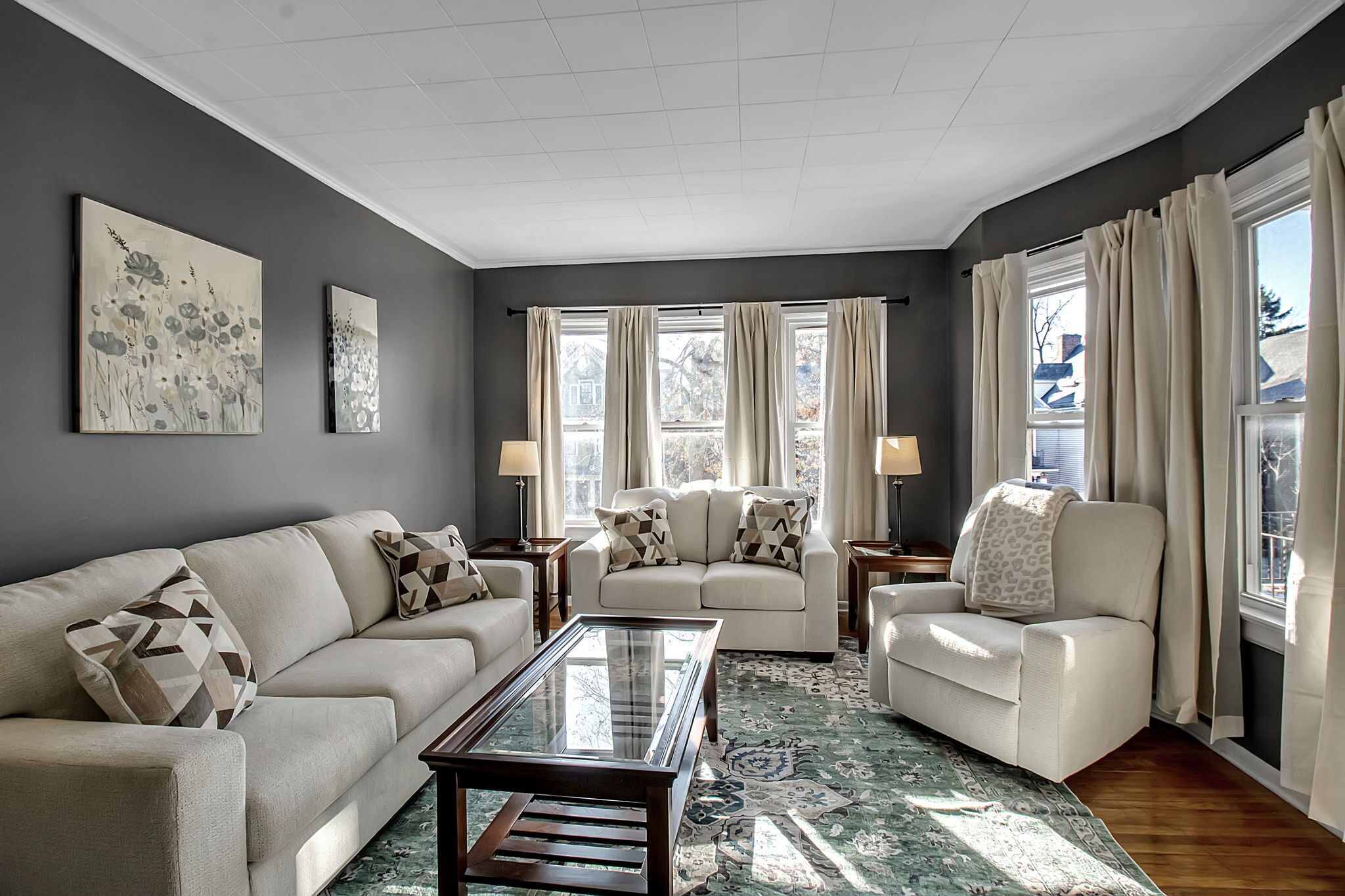Un soggiorno con un divano color beige, una poltrona abbinata e un tavolino in vetro. Due finestre con tende, un quadro appeso al muro e un tappeto decorato.