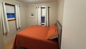A bedroom with a bed, two windows with curtains, a ceiling light, and a framed picture on the wall.