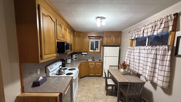 A compact kitchen with wooden cabinets, a white stove, a microwave, and a refrigerator. There is a dining table with chairs and a window with curtains.