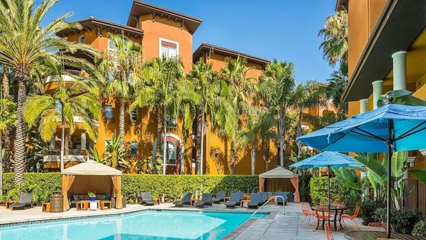 A swimming pool with lounge chairs and umbrellas, surrounded by palm trees and a building with balconies. - Charming Family Home | Heart of OC | Free Parking (Orange)