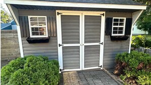 A small, single-story shed with a grey exterior and white trim. It features two windows with black shutters, a central door with a white frame, and a stone-paved entrance. - Litchfield Retreat, Wineries, Foliage & Skiing (Torrington)