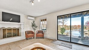 Een woonkamer met een open haard, twee leren fauteuils, een grote flatscreen-tv en uitzicht op een meer door middel van grote glazen deuren.