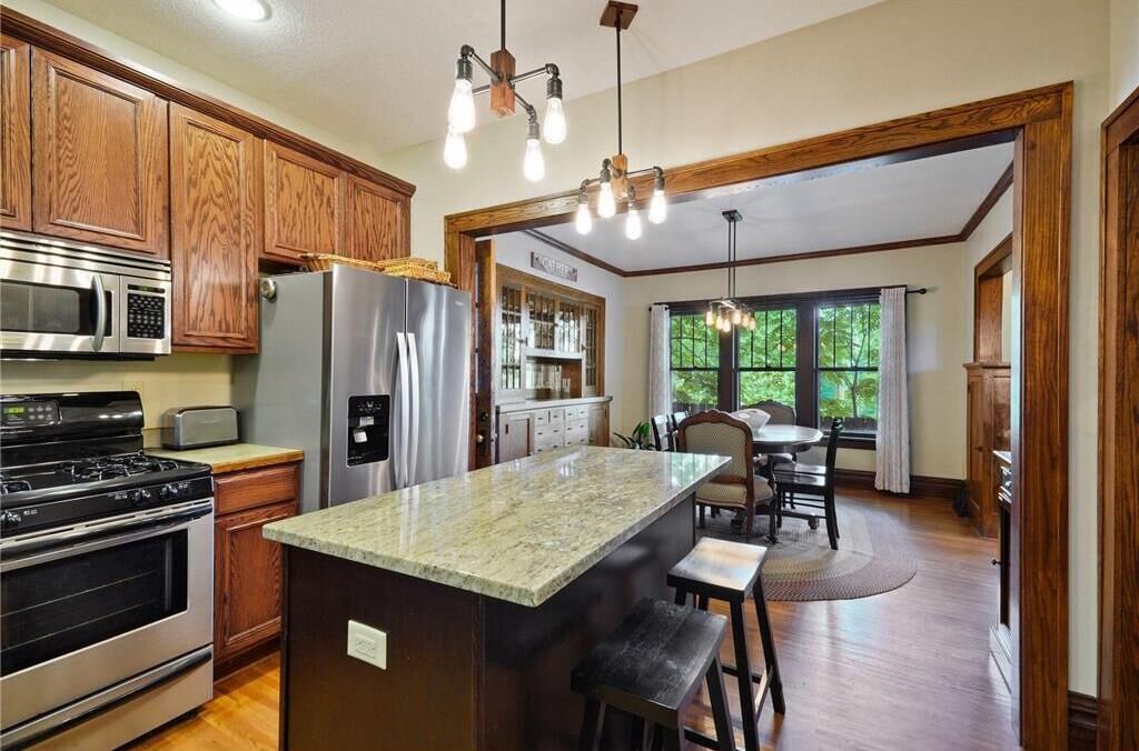 A kitchen with wooden cabinets, stainless steel appliances, a granite countertop island, and a dining area with a view of the outdoors.