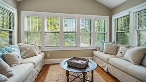Ein Wohnzimmer mit hoher Decke, großen Fenstern mit Jalousien, einem beigefarbenen Sofa, einem runden Couchtisch und einem Teppich.