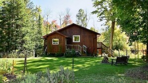 En stuga byggd av trä med en veranda, omgiven av en välskött gräsmatta och träd.