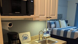 Une petite cuisine équipée d’un four à micro-ondes, d’une machine à café et d’un évier. On aperçoit un lit recouvert d’une couverture à rayures bleues et blanches au fond.