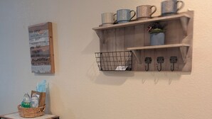 Un mur orné d’une étagère en bois où sont posées des tasses et une petite plante en pot, un panier en métal contenant un autre panier en osier, le tout placé sur une table, ainsi qu’une enseigne fixée au mur avec du texte.