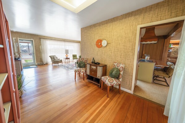 A spacious living room with wooden flooring, a large window, and a dining area with patterned chairs.