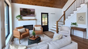 Un vaste salon doté d’un haut plafond en bois, d’un grand canapé gris, d’un fauteuil marron et d’une petite table basse agrémentée d’une plante. Un escalier avec des rampes en bois mène à l’étage supérieur.