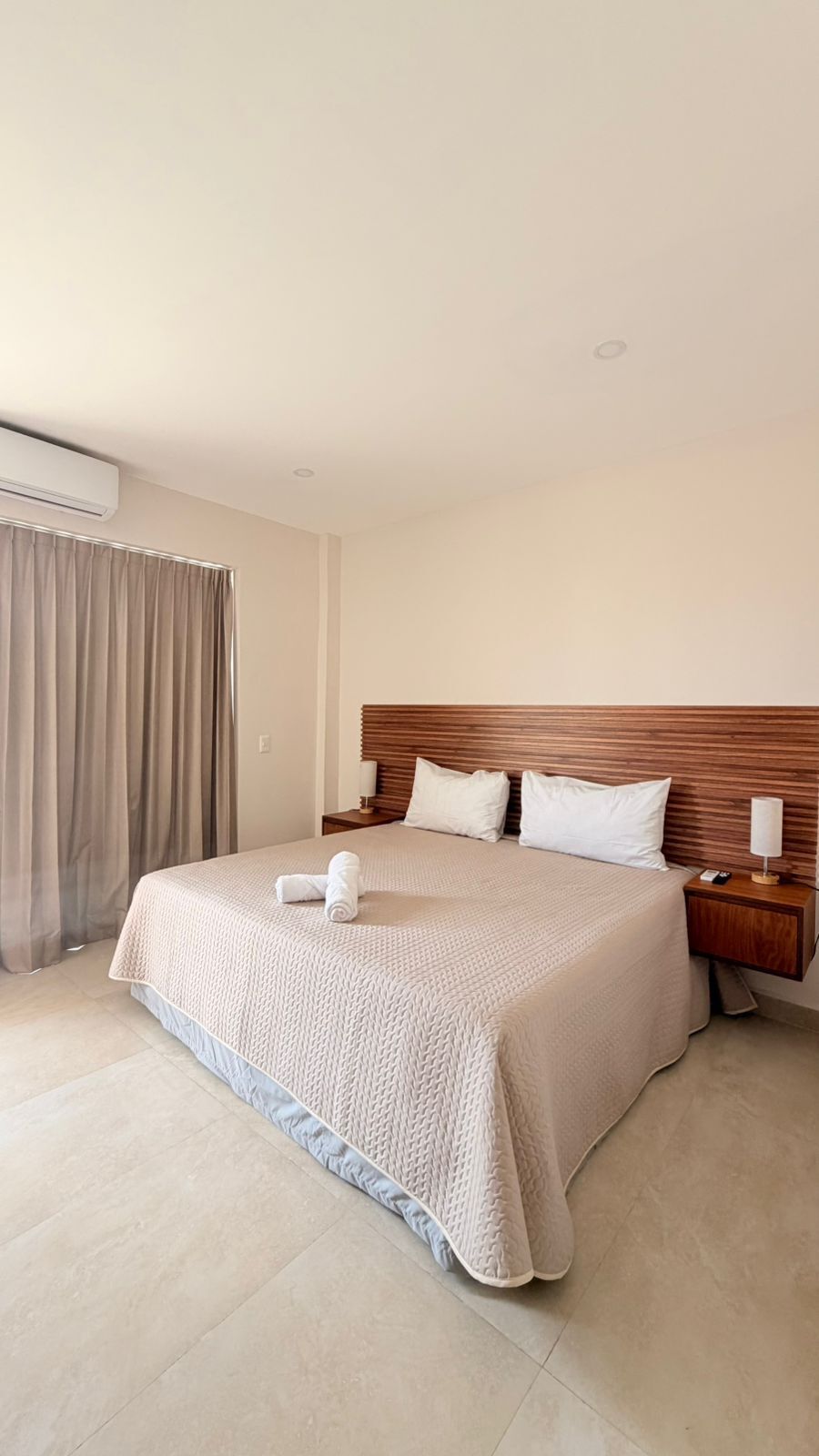 A hotel room with a large bed, wooden headboard, two bedside tables, and a window with curtains.