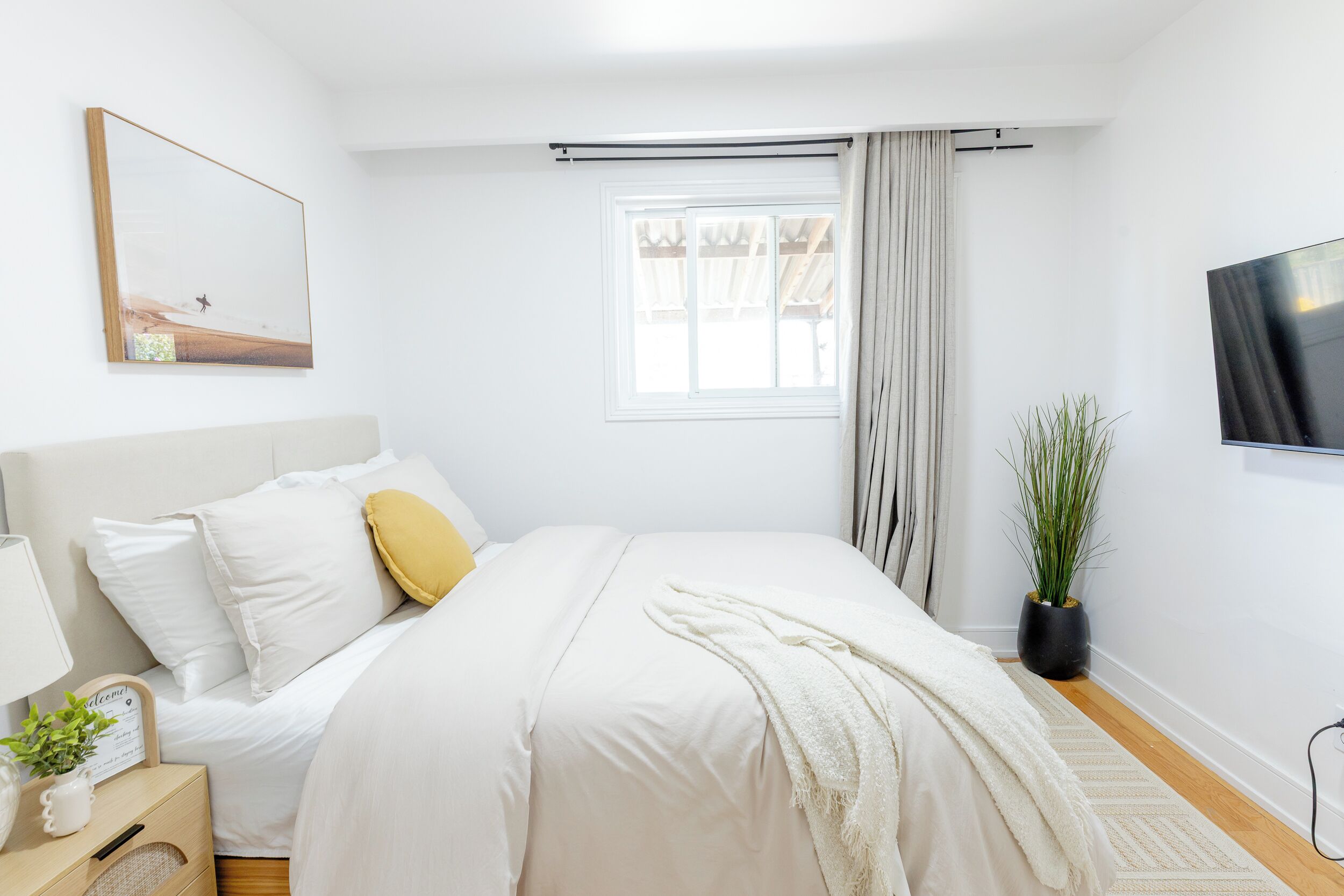 A bedroom with a bed, a bedside table with a lamp, a potted plant, a window with curtains, and a wall-mounted TV.