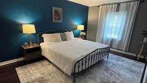 Une chambre à coucher comprenant un lit, deux tables de chevet avec des lampes, un luminaire au plafond, une fenêtre avec des rideaux et une bouche d’aération fixée au mur.