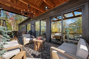 A covered outdoor patio with a fire pit, seating, and a view of the surrounding trees. - Highland House - Hot tub, fire pit, close to ski resorts (Banner Elk)