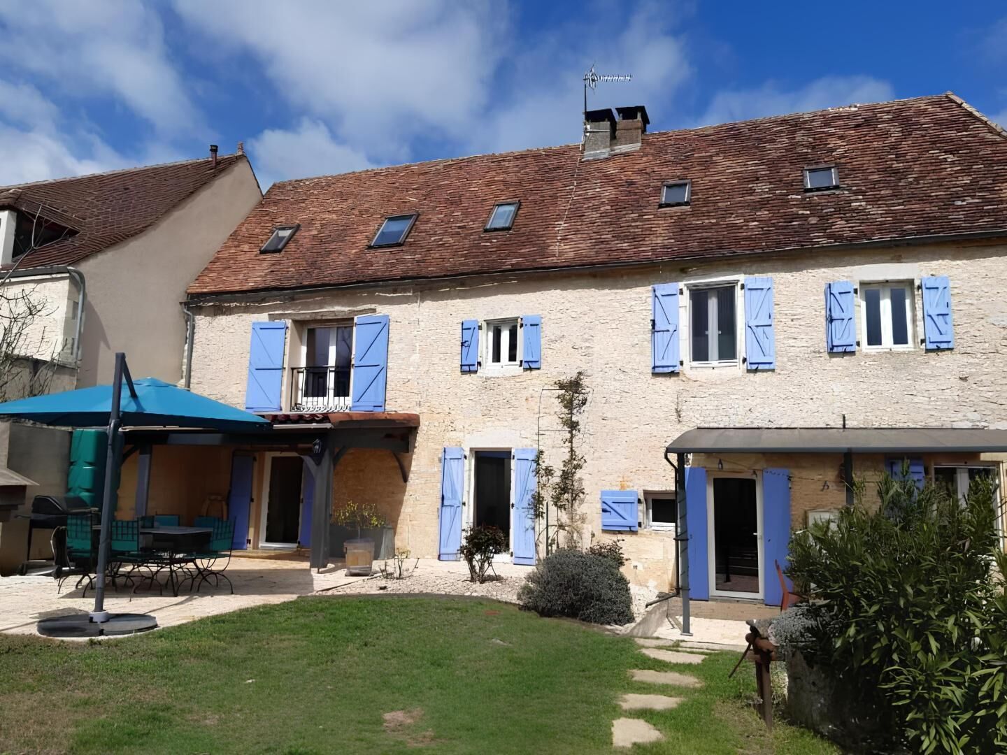 A two-story stone building with blue shutters, a covered patio with outdoor furniture, and a small garden area.