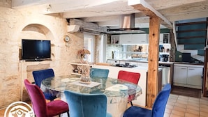 A dining area with a glass table surrounded by colorful chairs, a television on the wall, and a rustic kitchen in the background.