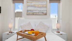 Una cama bien hecha con una bandeja de madera que sostiene frutas y un vaso de zumo de naranja, flanqueada por dos mesitas de noche con lámparas, en una habitación con un gran ventanal y un cuadro en la pared.