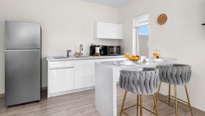 Una cocina moderna con refrigerador de acero inoxidable, muebles blancos, microondas integrado y una barra desayunadora con dos taburetes grises tapizados.