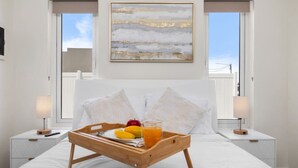 Una cama bien hecha con una bandeja de madera que contiene frutas y un vaso de jugo de naranja, flanqueada por dos mesitas de noche con lámparas, en una habitación con un ventanal grande y un cuadro sobre la cama.