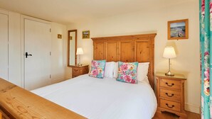 Un dormitorio con cama de madera, ropa de cama blanca, dos almohadas florales, una mesita de noche de madera con una lámpara y un espejo en la pared.