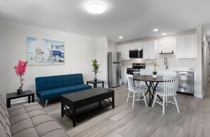 Uma sala de estar moderna com um sofá azul, uma mesa de jantar com cadeiras brancas, uma cozinha com armários brancos e um quadro com uma cena de praia.