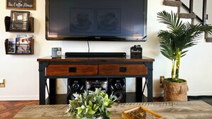 A living room with a flat-screen TV mounted on the wall, a wooden entertainment center with speakers, a potted plant, and a welcome sign. - Cozy Elevated Luxury - King Bed, Coffee Bar, Sleeps 5 (Springfield)