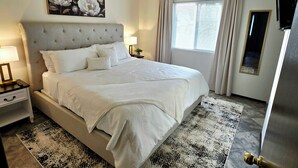 A neatly made bed with a tufted headboard, a bedside table with a lamp, a framed floral artwork above the bed, and a window with curtains. - Cozy Elevated Luxury - King Bed, Coffee Bar, Sleeps 5 (Springfield)