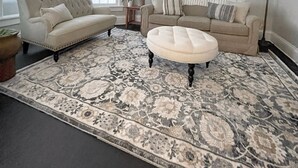 Uma sala de estar com um sofá bege, um banquinho combinando e um tapete estampado. Há três quadros na parede e um abajur.